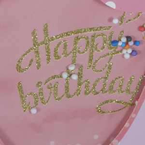 Detale aplikacji 3D w kształcie balonu na torebce. Złoty, brokatowy napis Happy Birthday oraz dekoracyjne, drobne kuleczki na różowym tle.