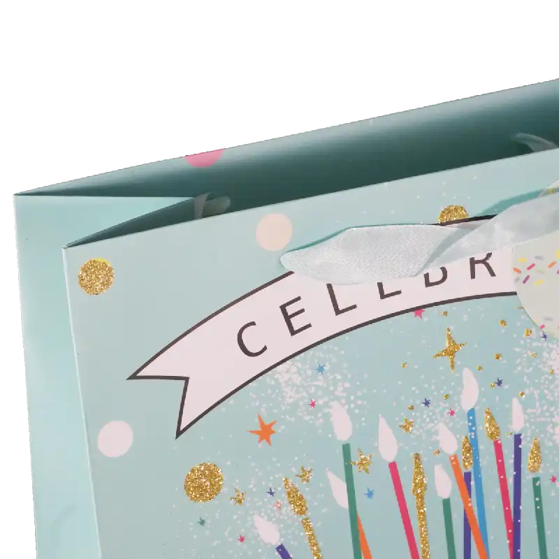 Zbliżenie na górną część torebki z napisem Celebrate, brokatowymi detalami oraz wstążkowym uchwytem.