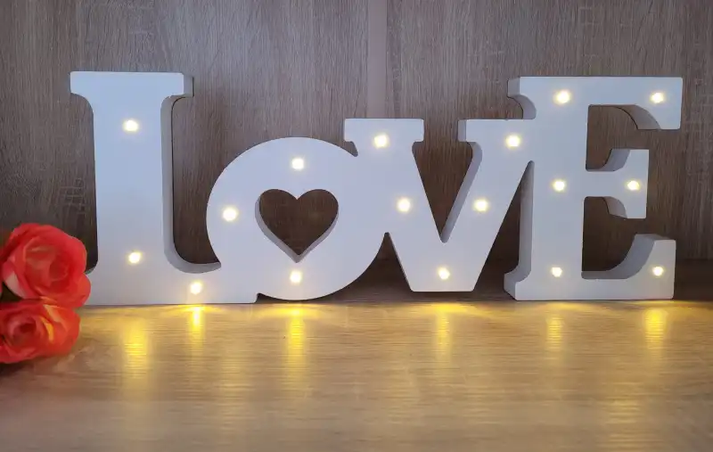 Drewniany napis LOVE 44 cm w białym kolorze, ustawiony na blacie obok dekoracyjnych różowych kwiatów. Nastrojowe oświetlenie LED tworzy romantyczny klimat w pomieszczeniu.