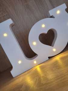 Zbliżenie na lewą stronę białego napisu LOVE 44 cm. Widoczny detal w kształcie serca wewnątrz litery O oraz ciepłe światło diod LED wkomponowanych w drewnianą konstrukcję.