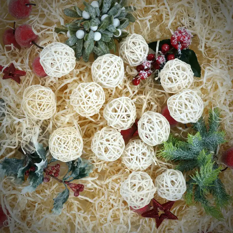 Artykuły dekoracyjne w postaci ażurowych kul wiklinowych z brokatem, ułożone w kompozycji wraz z gałązkami jemioły oraz czerwonymi owocami. Naturalne kule i gwiazdy rattanowe w typowo świątecznym otoczeniu pełnym ciepłych barw.