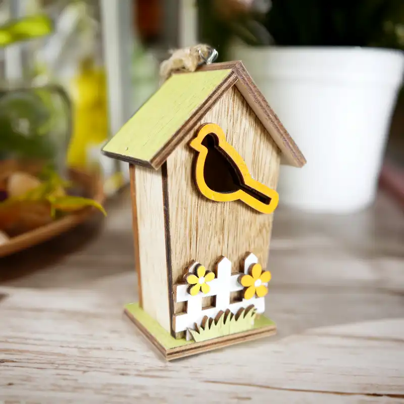 Drewniany domek zawieszka 10 cm z naturalnego drewna, zdobiony motywem ptaka oraz płotu z kwiatami. Ozdoba z jutowym sznurkiem w ujęciu z góry na stół.