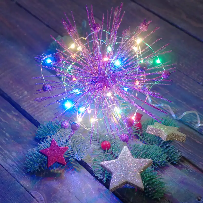 Świąteczna aranżacja z fioletowym jeżykiem 15 cm, który został ozdobiony światełkami LED. Dekoracja leży na gałązkach igliwia w towarzystwie błyszczących gwiazdek na drewnianym stole.
