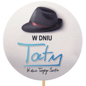 Zbliżenie na okrągły topper W Dniu Taty o średnicy 9 cm. Grafika przedstawia czarny kapelusz i niebieski napis na jasnym tle. Element dekoracyjny na długim piku.