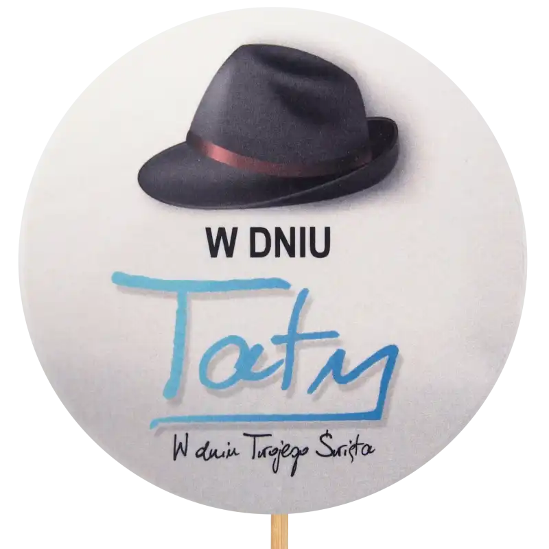 Zbliżenie na okrągły topper W Dniu Taty o średnicy 9 cm. Grafika przedstawia czarny kapelusz i niebieski napis na jasnym tle. Element dekoracyjny na długim piku.