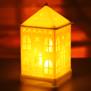 Zbliżenie na lampion LED w kształcie domku, który rozświetla ciemne tło ciepłym blaskiem, podkreślając przytulny charakter tej dekoracji.