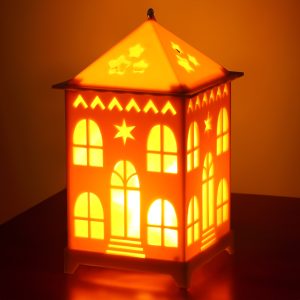 Zbliżenie na włączony lampion LED w kształcie domku na ciemnym tle. Intensywne ciepłe światło podkreśla wzory gwiazdek na dachu oraz ażurowe elementy ścianek.