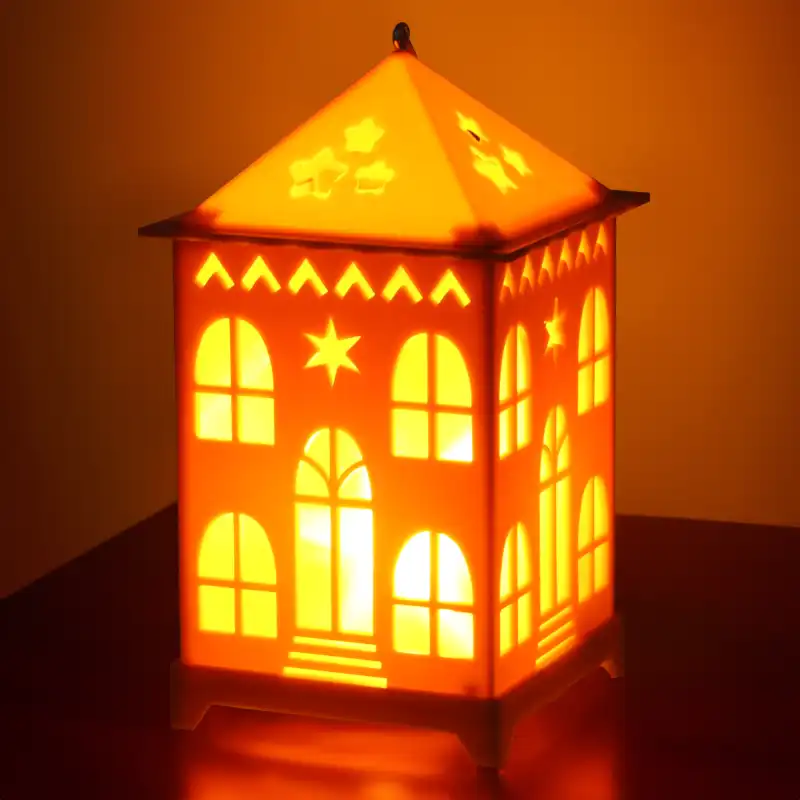 Zbliżenie na włączony lampion LED w kształcie domku na ciemnym tle. Intensywne ciepłe światło podkreśla wzory gwiazdek na dachu oraz ażurowe elementy ścianek.