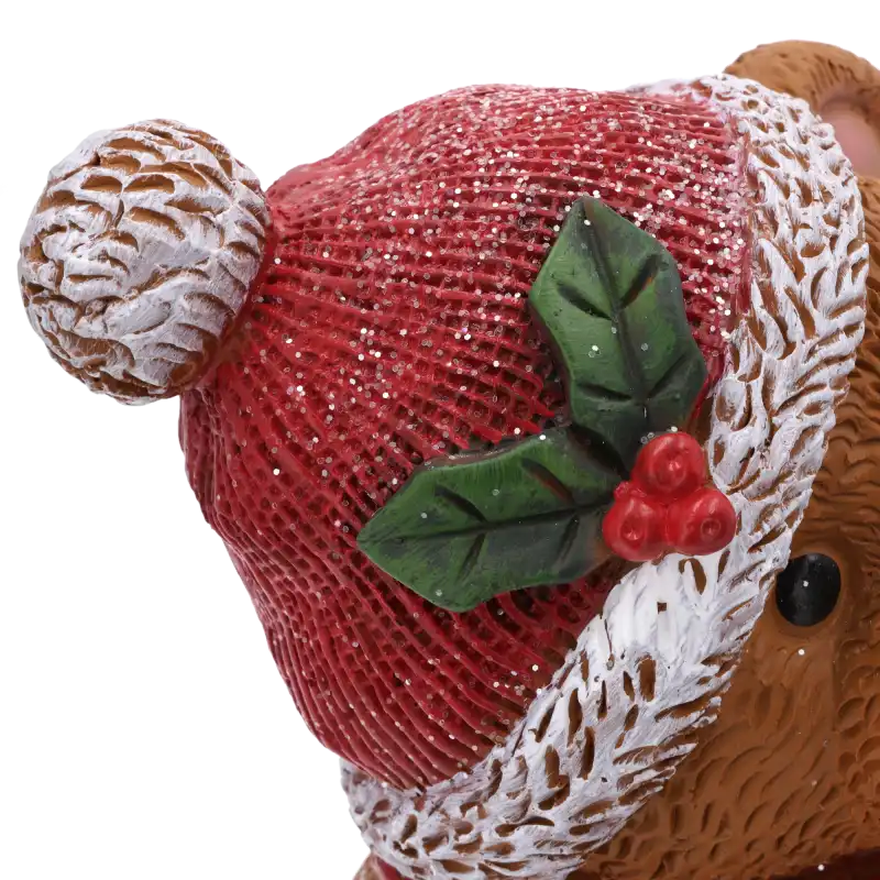 Zbliżenie na czerwoną czapkę misia z pomponem. Widoczne zdobienia w postaci zielonych liści i czerwonych jagód oraz warstwa brokatu, która dodaje figurce blasku.