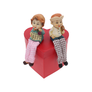 Figurki babci i dziadka siedzące na czerwonym pudełku w kształcie serca. Dekoracja podkreśla urokliwy design postaci ze zwisającymi nogami.
