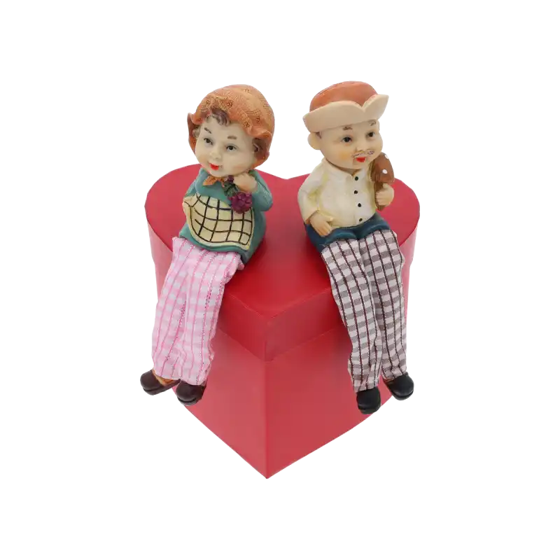 Figurki babci i dziadka siedzące na czerwonym pudełku w kształcie serca. Dekoracja podkreśla urokliwy design postaci ze zwisającymi nogami.