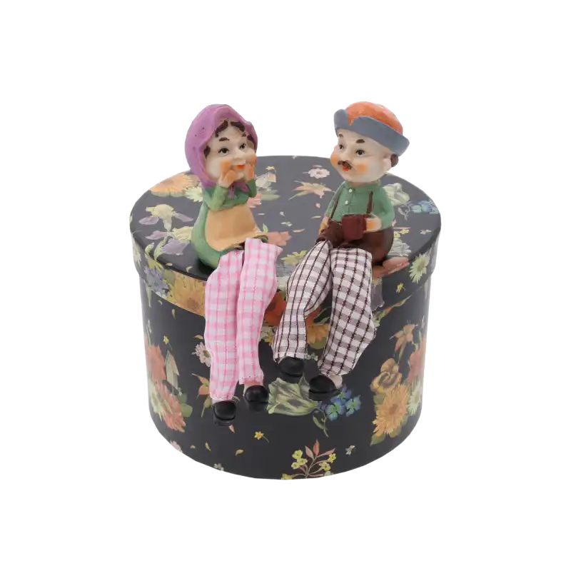 Figurka dziadka obok figurki babci, siedzące na ozdobnym okrągłym pudełku. Kompozycja dekoracyjna prezentująca obie postacie.