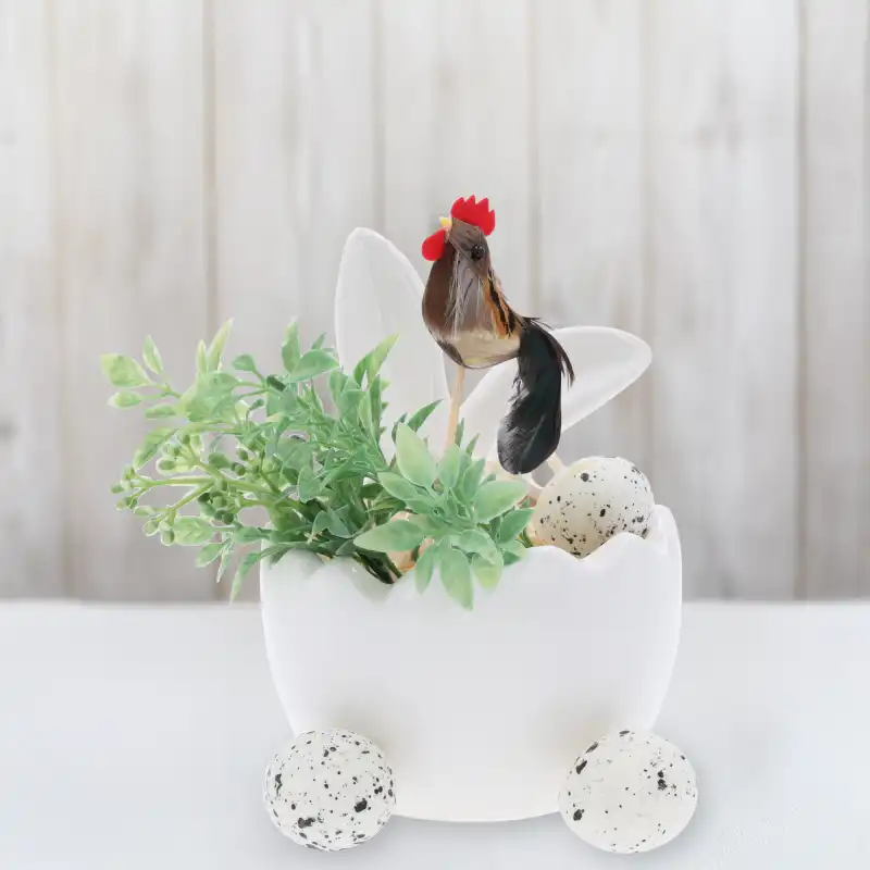 Aranżacja wielkanocna z kogutem na piku, jajkiem ceramicznym i roślinnością na jasnym, drewnianym tle w rustykalnym stylu.
