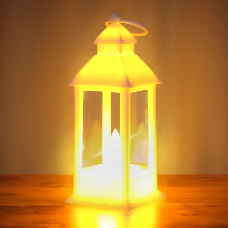 Biały lampion LED 15 cm emitujący ciepłe, nastrojowe światło. Dekoracja ustawiona na drewnianym blacie, ukazująca efekt świecenia w przyciemnionym otoczeniu.