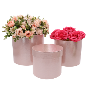 Dwa jasnoróżowe flower boxy wypełnione sztucznymi kwiatami w odcieniach różu, stojące obok pustego pudełka z pokrywką.