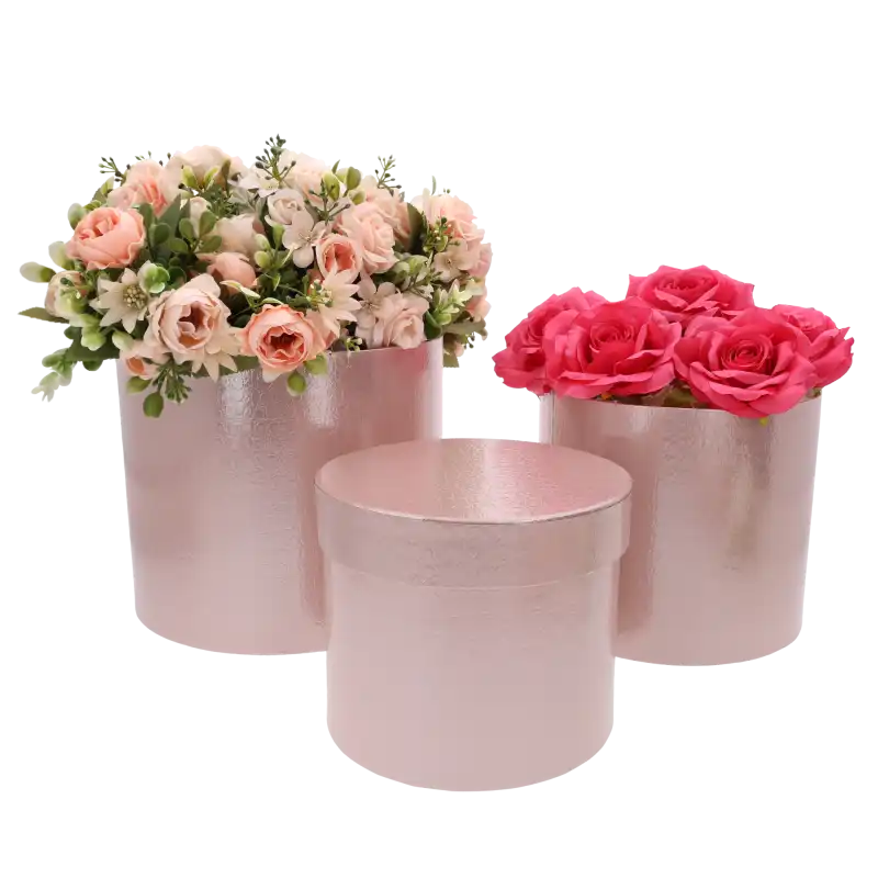 Dwa jasnoróżowe flower boxy wypełnione sztucznymi kwiatami w odcieniach różu, stojące obok pustego pudełka z pokrywką.