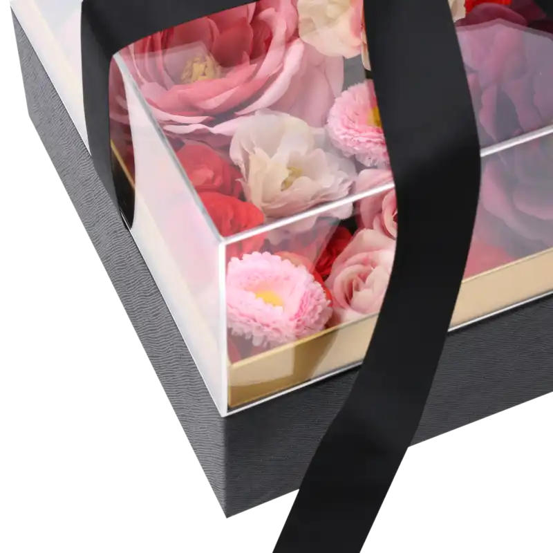 Zbliżenie na wnętrze wypełnionego kwiatami czarnego flower boxa pod przezroczystą pokrywką z przewiązaną czarną wstążką.