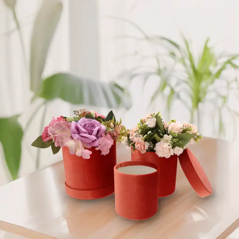 Aranżacja trzech welurowych flower boxów w kolorze rudym na jasnym blacie, dwa z nich udekorowane kompozycjami kwiatowymi.