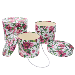 Trzy okrągłe pudełka z nadrukiem roślinnym, z których jedno jest puste i otwarte, a drugie ma położoną obok pokrywkę. W tle trzeci flower box.