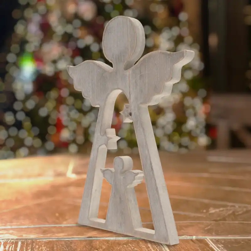 Drewniana figurka anioła 30 cm ustawiona na drewnianym podłożu. W tle widoczny rozmyty zarys świątecznej choinki z kolorowymi lampkami.