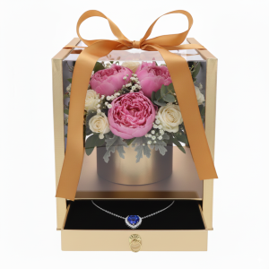 Widok na złoty flower box z przezroczystymi ściankami. W środku bukiet róż i piwonii, a w wysuniętej szufladzie naszyjnik z niebieskim kamieniem.