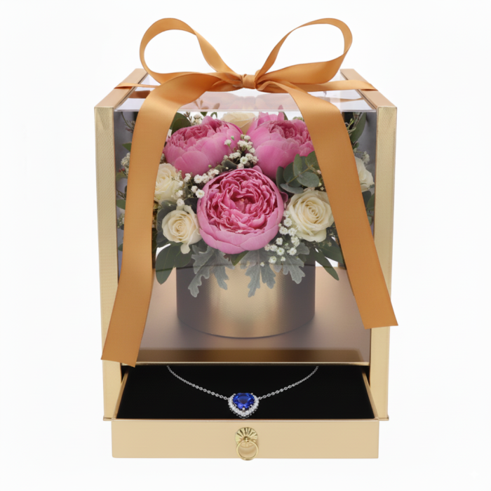 Widok na złoty flower box z przezroczystymi ściankami. W środku bukiet róż i piwonii, a w wysuniętej szufladzie naszyjnik z niebieskim kamieniem.