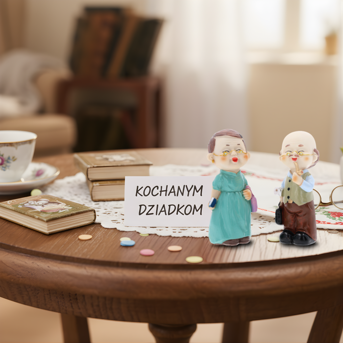 Figurki babcia i dziadek ustawione na drewnianym stole obok wizytówki z napisem Kochanym Dziadkom, tworzące klimatyczną aranżację prezentową.