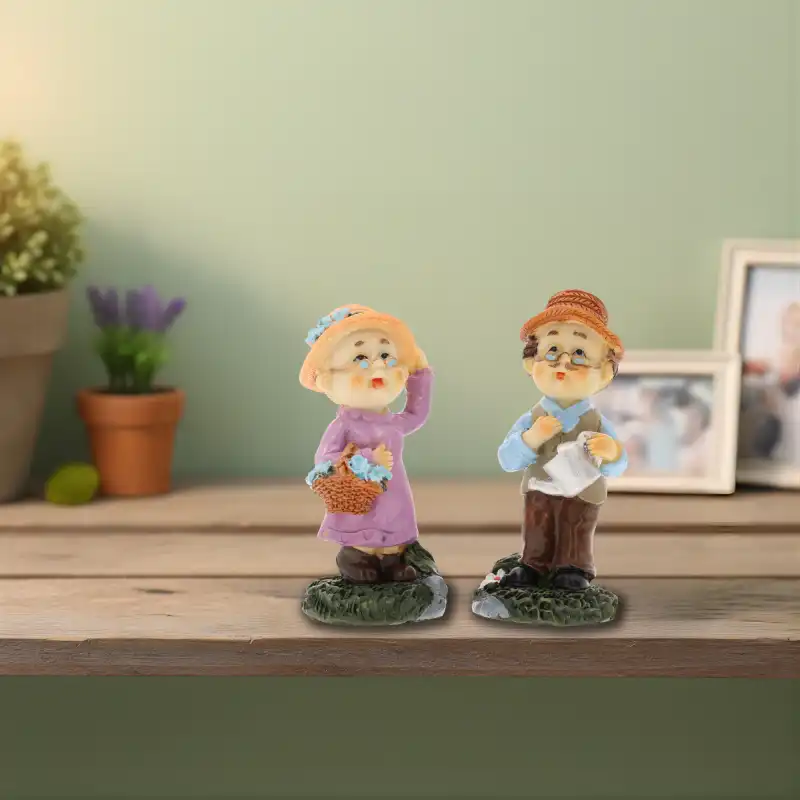 Figurki babcia i dziadek ustawione na drewnianym blacie w tle z roślinami doniczkowymi i ramkami na zdjęcia, tworząc domowy klimat.