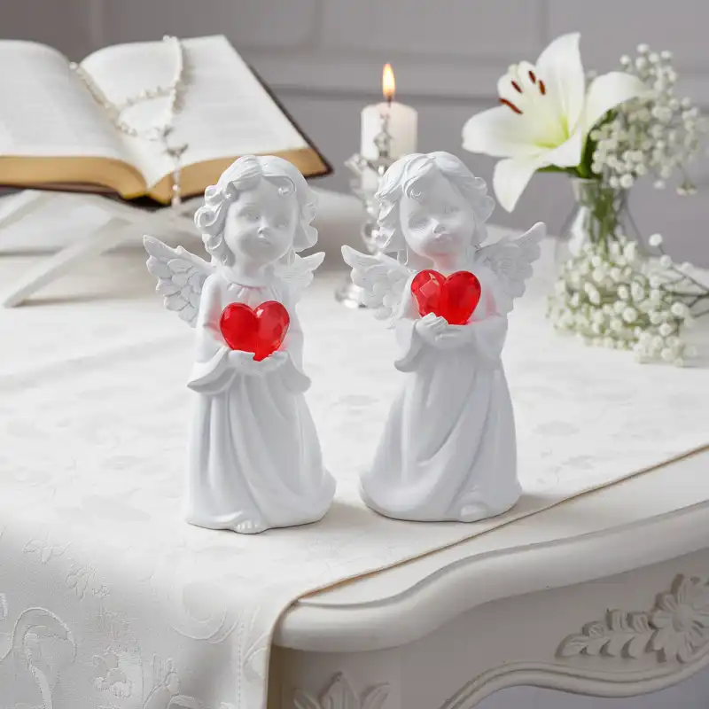 Dwie figurki aniołków z czerwonymi sercami ustawione obok siebie na jasnym obrusie w otoczeniu kwiatów i świecy.