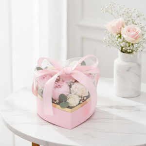 Różowy flower box w kształcie serca ustawiony na białym stole w jasnym pomieszczeniu, z wazonem kwiatów w tle.