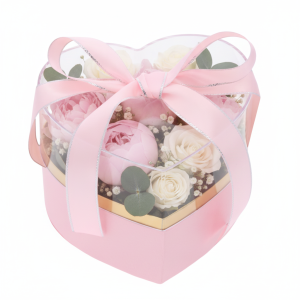 Różowy flower box serce 15x14x12 cm z przezroczystą pokrywką, złotym wnętrzem i przewiązany różową wstążką, wypełniony kwiatami.