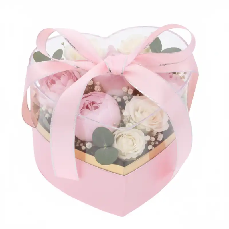 Różowy flower box serce 15x14x12 cm z przezroczystą pokrywką, złotym wnętrzem i przewiązany różową wstążką, wypełniony kwiatami.