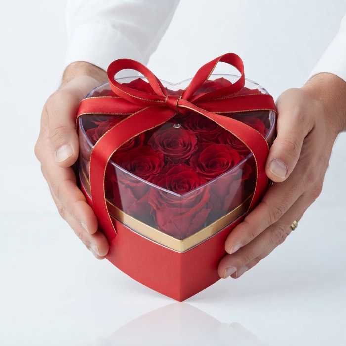 Czerwony flower box w kształcie serca ze złotym wnętrzem, przezroczystą pokrywką i satynową wstążką, trzymany w dłoniach jako eleganckie pudełko prezentowe.
