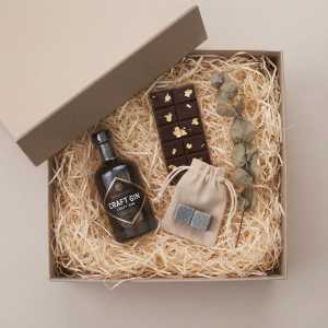 Eleganckie pudełko prezentowe wypełnione wełną drzewną, zawierające butelkę, czekoladę oraz zestaw kamieni w woreczku. Idealny pomysł na upominek.