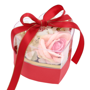 Czerwony flower box z kompozycją kwiatową wewnątrz, zabezpieczony przezroczystą pokrywką z czerwoną kokardą.