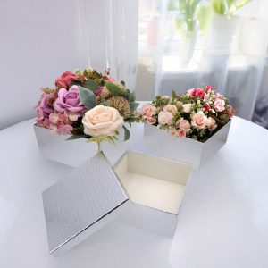 Srebrne pudełka typu flower boxy wypełnione kwiatami, ustawione na białym blacie. Widoczne jedno puste pudełko z pokrywką w układzie pozwalającym na prezentację wnętrza.