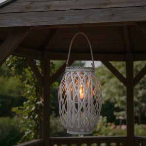 Lampion wiklinowy zawieszony w altanie, z zapaloną świecą wewnątrz szklanego wkładu, tworzy nastrojowe oświetlenie w ogrodzie.