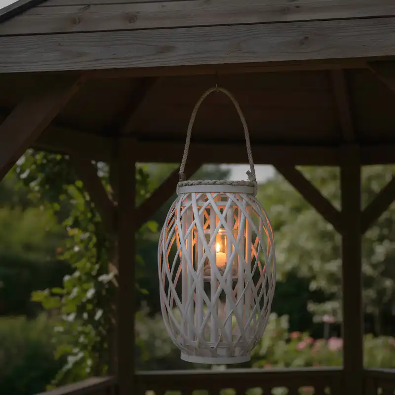 Lampion wiklinowy zawieszony w altanie, z zapaloną świecą wewnątrz szklanego wkładu, tworzy nastrojowe oświetlenie w ogrodzie.