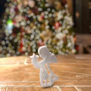 Figurka anioła z drewna o wysokości 14 cm postawiona na drewnianym blacie. W tle rozmyte świąteczne oświetlenie choinkowe tworzące przytulny klimat.