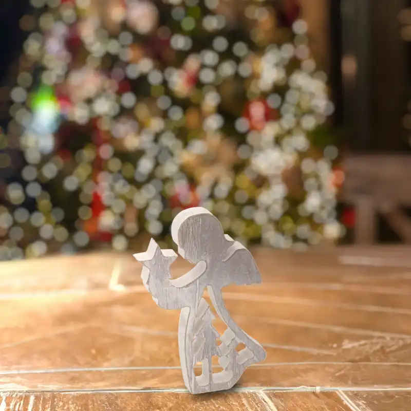 Figurka anioła z drewna o wysokości 14 cm postawiona na drewnianym blacie. W tle rozmyte świąteczne oświetlenie choinkowe tworzące przytulny klimat.