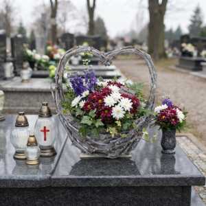 Kwiatowa dekoracja nagrobna w koszyku z gałęzi w kształcie serca, ustawiona na płycie pomnika obok zniczy.
